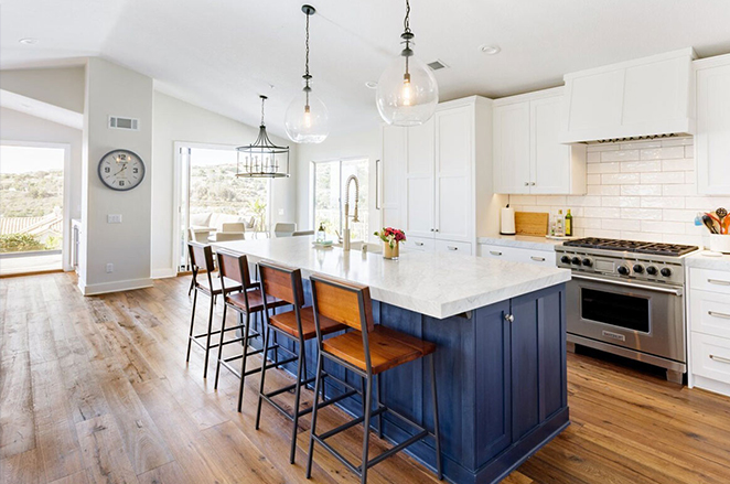 Bright, modern kitchen with a large white island featuring a navy base, four wooden barstools, stainless steel appliances, white cabinets, pendant lights, and wooden flooring. Sunlight streams through large windows.