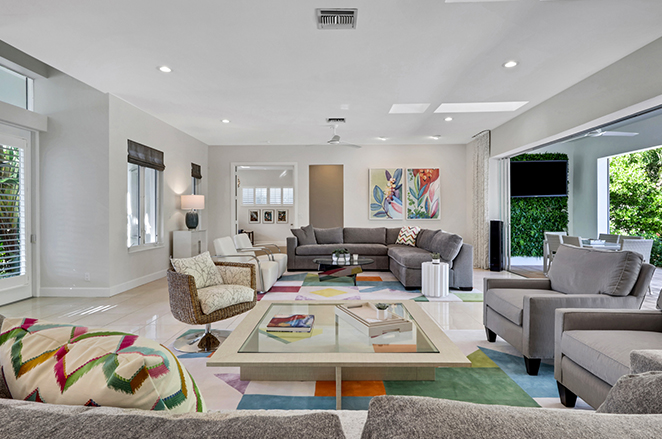 Contemporary living room with gray sofas, armchairs, and a glass coffee table on a colorful geometric rug. Large windows and sliding doors let in natural light, and vibrant artwork decorates the walls.
