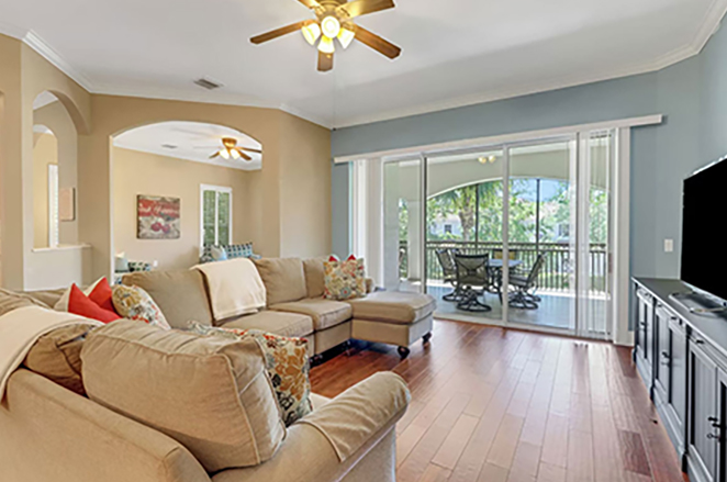 Bright living room with a sectional sofa, colorful pillows, ceiling fan, wooden floors, TV stand, and large sliding glass doors opening to a covered patio with outdoor seating and greenery visible outside.