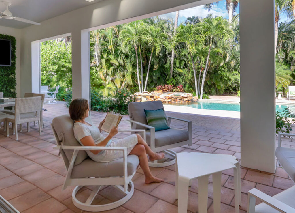 A woman sits in a chair on a covered patio, reading a book. The patio overlooks a pool surrounded by tall palm trees and lush greenery on a sunny day. There are tables and chairs on the patio.
