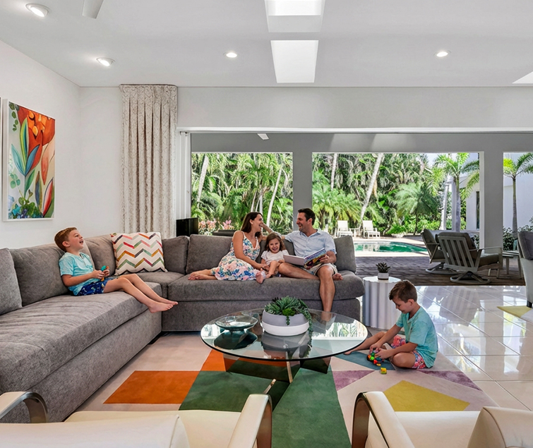 A family of four relaxes in a bright, modern living room with a large gray sectional. The parents and daughter sit together, while two sons play separately. Large windows reveal a lush tropical garden outside.