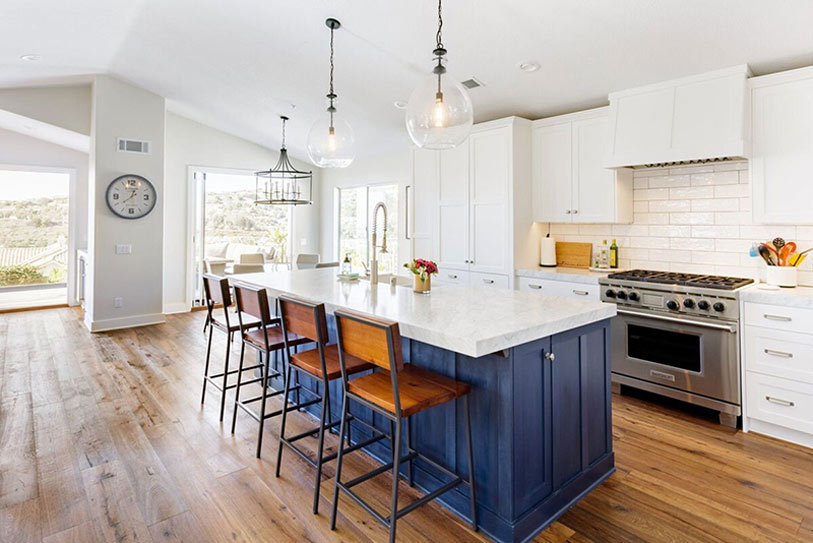 Modern kitchen with a large marble island, blue base cabinets, four wooden bar stools, stainless steel appliances, white cabinetry, pendant lights, and wide plank wood flooring, with sunlight streaming in.