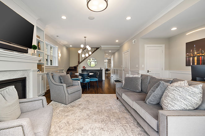 Modern living room with gray sofas, armchairs, a large rug, wall-mounted TV above a fireplace, dining table, and staircase leading upstairs. Space is bright with recessed lighting and neutral-colored walls.